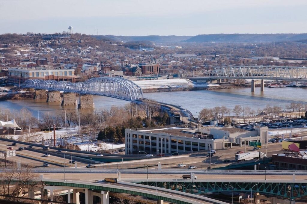 A sweeping winter view from Cincinnati's Eden Park looking across the Ohio River to the city of Newport, Kentucky. The image captures the bustling infrastructure, including the distinctive blue arched bridge, that connects these two cities, highlighting Newport's prime location and its scenic riverfront setting.